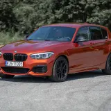 Pomarańczowe BMW 140i z czarnymi felgami stoi na asfalcie, w tle drzewa.