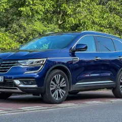 Granatowy Renault Koleos stoi na czerwonej kostce brukowej, otoczony zielenią.