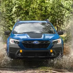 Niebieski Subaru Outback Wilderness z impetem wjeżdża w kałużę, tworząc fontannę wody i błota.