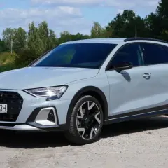 Jasnoszary Audi A3 Allstreet z czarnymi dodatkami stoi na żwirowej drodze nad jeziorem.