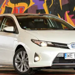 Biała Toyota Auris 2015 z polskimi tablicami rejestracyjnymi WE 974EU zaparkowana przed kolorowym muralem.