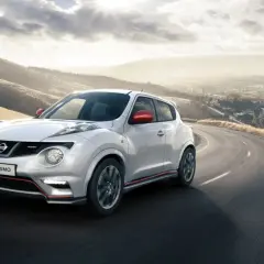 Biały Nissan Juke Nismo z czerwonymi lusterkami pędzi po krętej drodze z widokiem na miasto w oddali.
