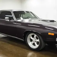 Klasyczny Plymouth Barracuda w ciemnym kolorze, z chromowanymi felgami i sportowym wlotem powietrza na masce.