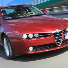 Czerwone Alfa Romeo 159 1.9 JTD pędzi po drodze, kierowca skupiony na trasie.