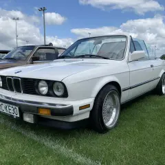 Białe BMW 325i kabriolet z chromowanymi felgami stoi na trawie pod błękitnym niebem z chmurami.