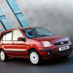 Czerwony Ford Fusion 1.4 benzyna w ruchu, z kobietą za kierownicą, na tle nowoczesnego budynku.