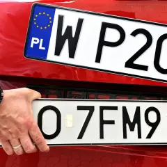 Ręka z bransoletką montuje tablicę rejestracyjną z napisem "O 7FM91" na czerwonym samochodzie. Powyżej widać inną tablicę "W P20".