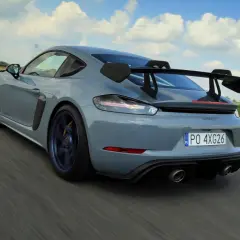 Szary Porsche GT4 RS z dużym skrzydłem pędzi po drodze, zostawiając za sobą rozmyte pola i błękitne niebo.