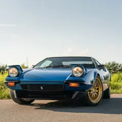Niebieski De Tomaso Pantera z wyskakującymi reflektorami i złotymi felgami na tle zieleni i błękitnego nieba.