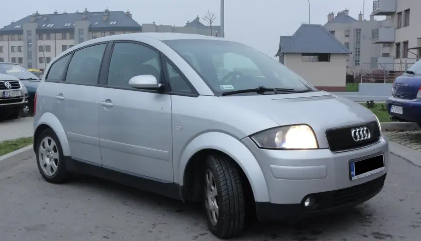Srebrne Audi A2 z włączonymi światłami, zaparkowane na tle nowoczesnych budynków mieszkalnych.
