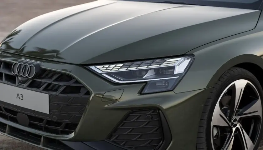 Nowe Audi A3 w zielonym kolorze prezentuje się elegancko. Widoczny fragment maski, grill z logo i reflektor podkreślają jego nowoczesny design.