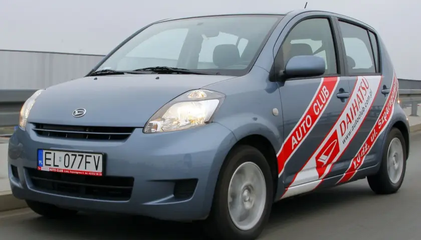 Szary Daihatsu Sirion z napisem "AUTO CLUB DAIHATSU" na boku, jadący po drodze.
