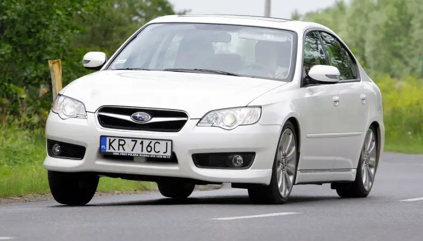 Białe Subaru Legacy z polskimi tablicami KR 716CJ jedzie po drodze wśród zieleni.