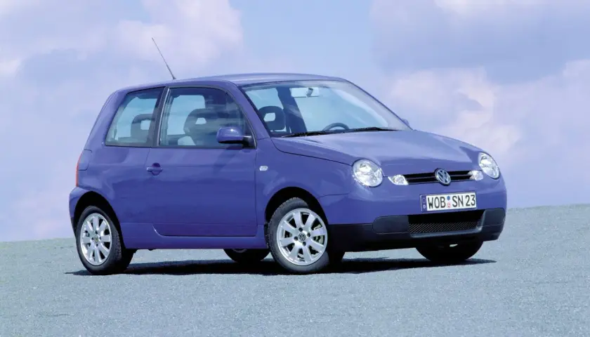 Fioletowy Volkswagen Lupo z tablicą rejestracyjną WOB SN 23 stoi na szarej nawierzchni pod błękitnym niebem z chmurami.