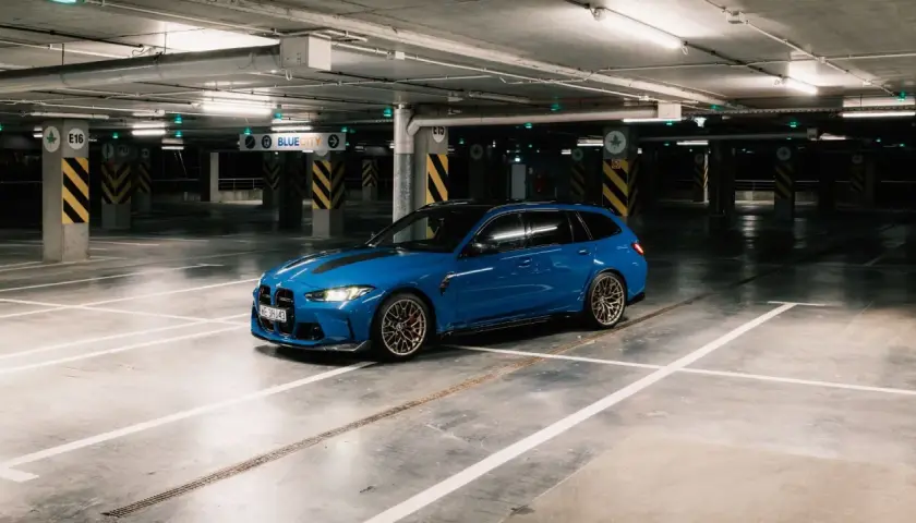Niebieskie BMW M3 Touring z czarnymi pasami na masce stoi na pustym parkingu podziemnym.