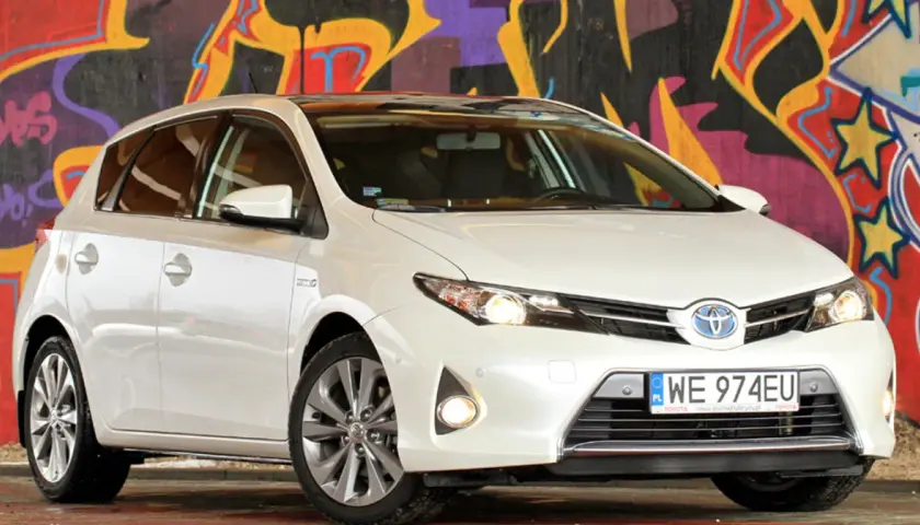 Biała Toyota Auris 2015 z polskimi tablicami rejestracyjnymi WE 974EU zaparkowana przed kolorowym muralem.