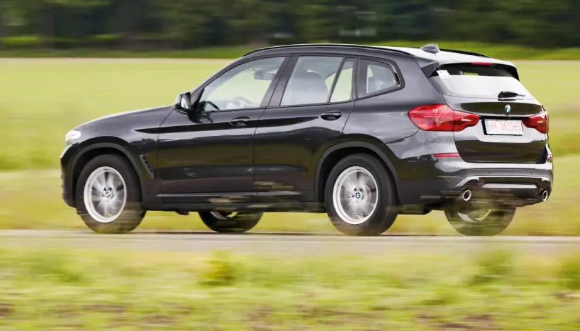 Ciemnoszary BMW G01 pędzi po wiejskiej drodze, rozmazując zielone pola i drzewa w tle.