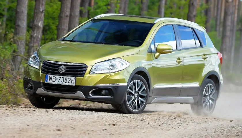 Zielony Suzuki S-Cross na drodze leśnej, wzbudzający pozytywne suzuki s-cross opinie. Kurz spod kół świadczy o jego terenowych możliwościach.