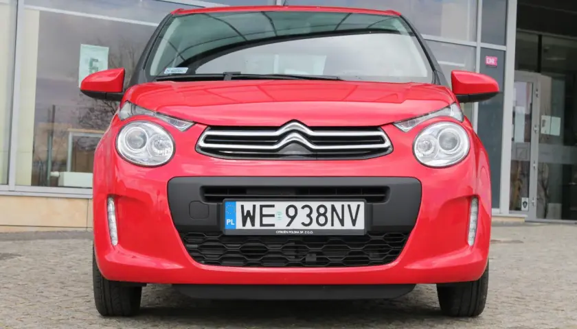 Czerwony Citroen C1 z tablicą rejestracyjną WE 938NV stoi przed budynkiem z dużymi oknami.