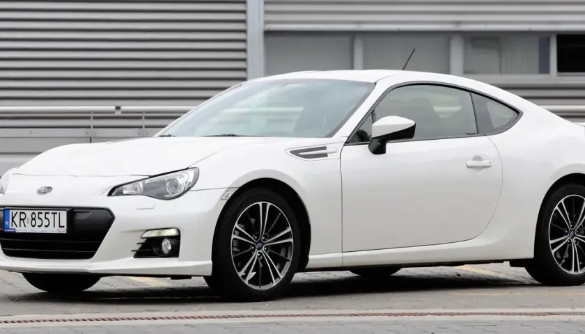 Białe Toyota BRZ z czarnymi felgami stoi przed budynkiem z poziomymi żaluzjami.