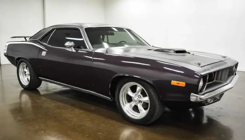 Klasyczny Plymouth Barracuda w ciemnym kolorze, z chromowanymi felgami i sportowym wlotem powietrza na masce.