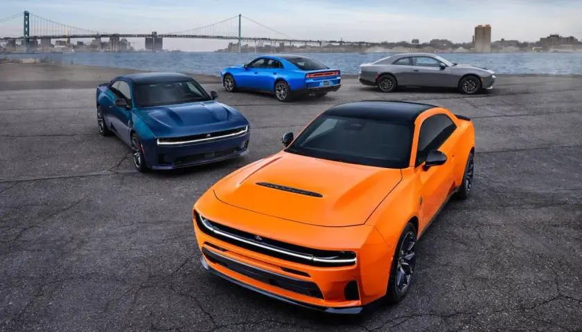 Pomarańczowy i niebieski Dodge Charger Hellcat oraz szary coupe stoją nad wodą z mostem w tle.