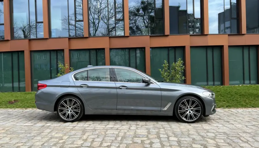 Szary sedan BMW M5 G30 stoi na brukowanym podjeździe przed nowoczesnym budynkiem z brązowymi ramami okien.