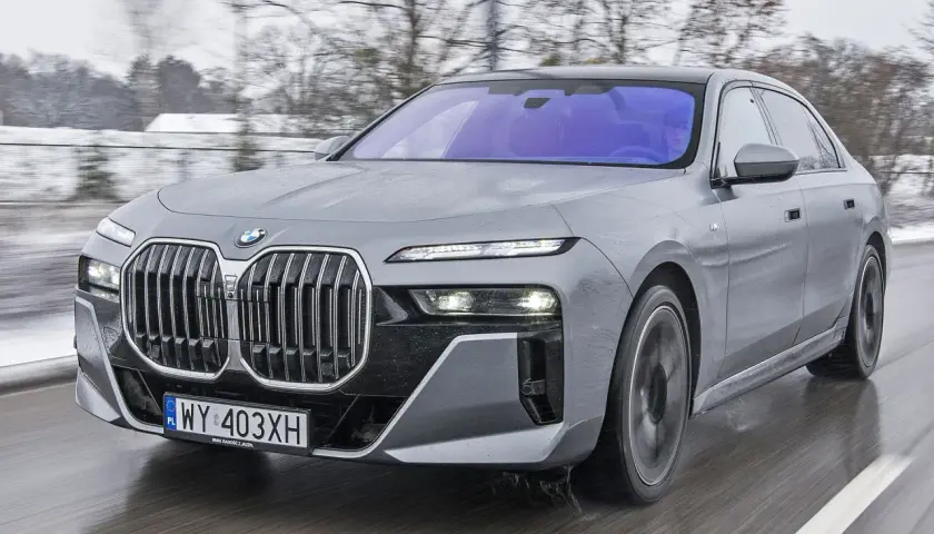 Szary BMW 7 2025 pędzi po mokrej drodze, ukazując swoją nowoczesną sylwetkę i charakterystyczny grill.