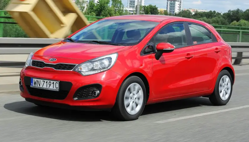 Czerwony Kia Rio 3 w ruchu, z widocznym numerem rejestracyjnym WN 7191C, na tle mostu i budynków.