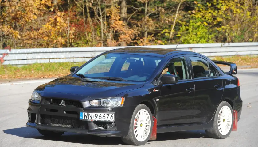 Czarny Mitsubishi Lancer 9 z białymi felgami i czerwonymi chlapacami na tle jesiennego lasu.