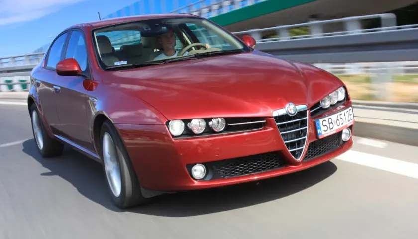 Czerwone Alfa Romeo 159 1.9 JTD pędzi po drodze, kierowca skupiony na trasie.