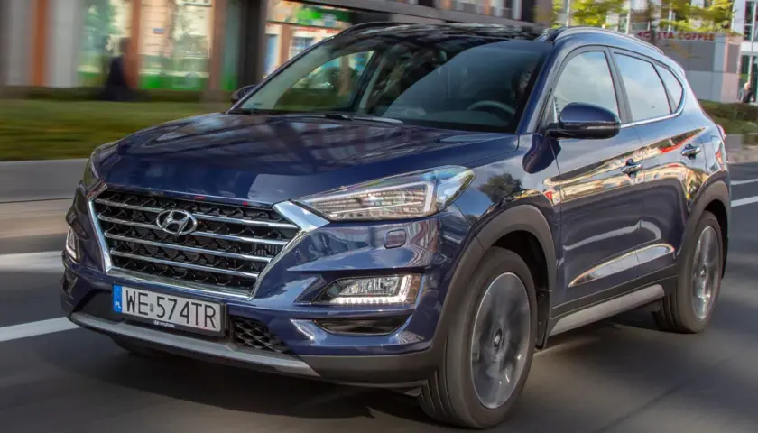 Nowy Hyundai Tucson w ciemnoniebieskim kolorze jedzie ulicą. Elegancka krata wlotu powietrza i nowoczesne reflektory podkreślają jego dynamiczny wygląd.