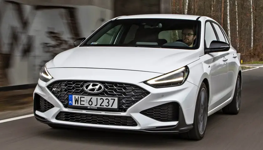 Biały Hyundai i30 N z dynamicznym grillem i agresywnym zderzakiem jedzie po drodze wśród drzew.