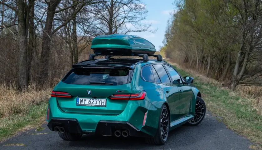 Zielone BMW M5 Touring z boxem dachowym na tle drzew i nieba.