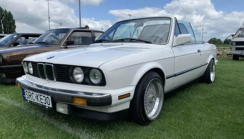 Białe BMW 325i kabriolet z chromowanymi felgami stoi na trawie pod błękitnym niebem z chmurami.