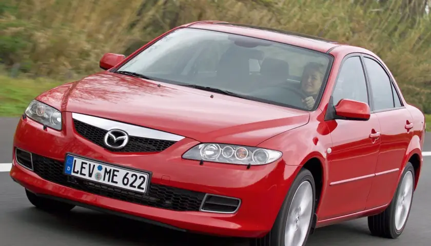 Czerwona Mazda 6 GG w ruchu, z uśmiechniętą kobietą za kierownicą, na tle zieleni.