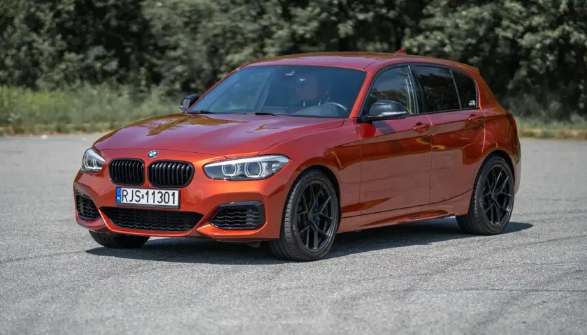 Pomarańczowe BMW 140i z czarnymi felgami stoi na asfalcie, w tle drzewa.