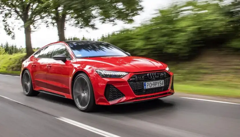Czerwone Audi RS7 pędzi po drodze, otoczone zielenią drzew.