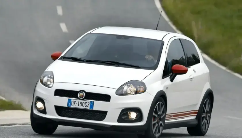 Biały Fiat Punto Evo Abarth z czerwonymi lusterkami pędzi po zakręcie na asfaltowej drodze.