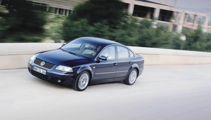 Ciemnoniebieski Volkswagen Passat B5 jedzie po moście, za nim rozmyte drzewa i budynek.