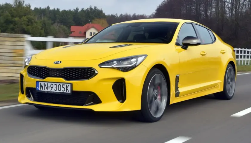 Żółty Kia Stinger GT pędzi po drodze, otoczony zielenią i domami.