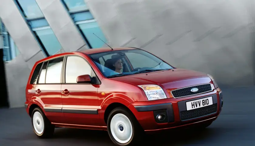 Czerwony Ford Fusion 1.4 benzyna w ruchu, z kobietą za kierownicą, na tle nowoczesnego budynku.