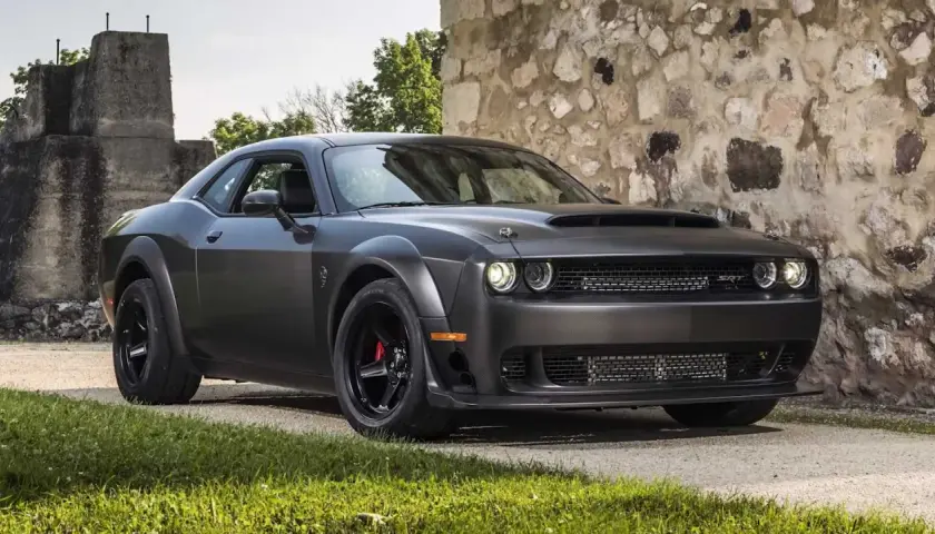 Mroczny Dodge Challenger Demon, z potężną maską i agresywnym grillem, stoi przed kamienną ścianą.