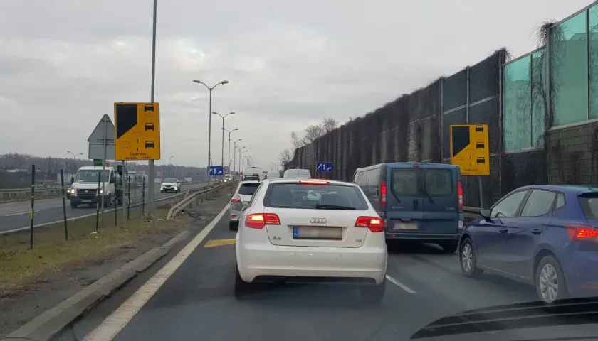 Korek na drodze, samochody stoją w miejscu. Widoczny znak "Zalecana zasada ruchu" informujący o konieczności stosowania jazdy na suwak.