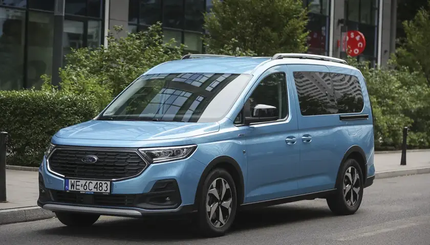 Błękitny ford 7 osobowy na tle nowoczesnego budynku. Idealny dla rodziny na wakacyjne wyprawy.