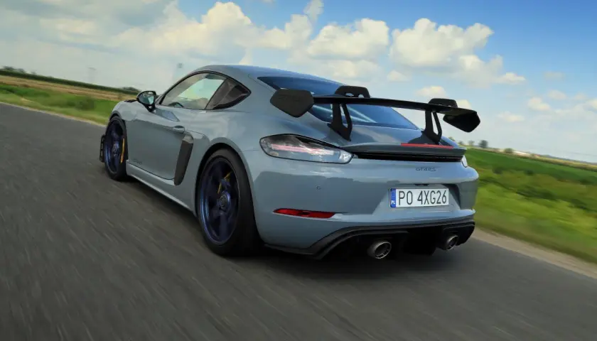 Szary Porsche GT4 RS z dużym skrzydłem pędzi po drodze, zostawiając za sobą rozmyte pola i błękitne niebo.