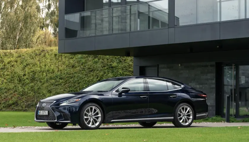 Elegancki, ciemnogranatowy Lexus LS 500 zaparkowany przed nowoczesnym domem z dużymi oknami i zielonym ogrodem.