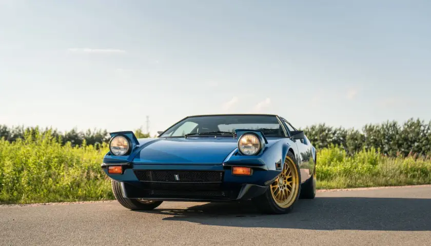Niebieski De Tomaso Pantera z wyskakującymi reflektorami i złotymi felgami na tle zieleni i błękitnego nieba.