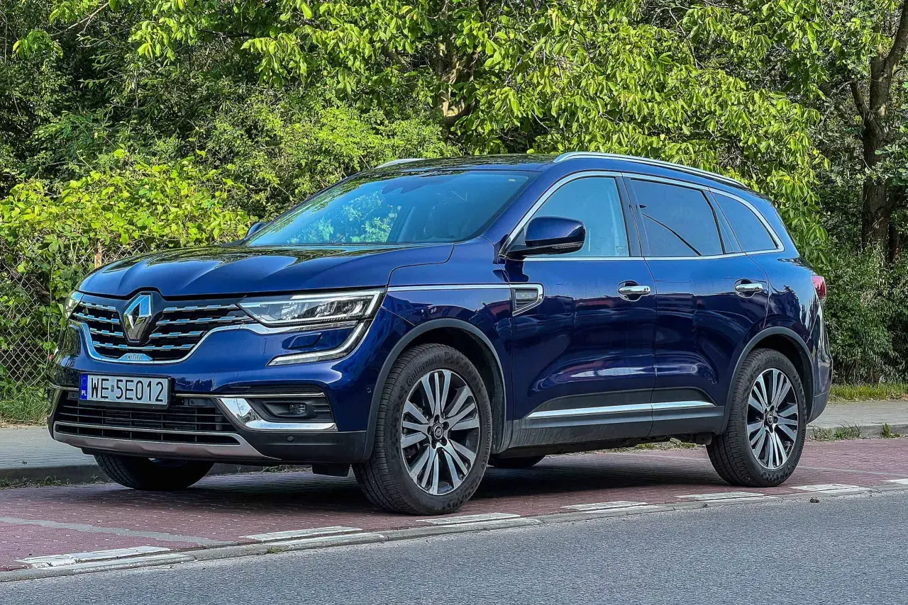 Granatowy Renault Koleos stoi na czerwonej kostce brukowej, otoczony zielenią.