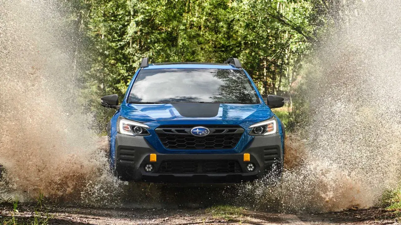 Niebieski Subaru Outback Wilderness z impetem wjeżdża w kałużę, tworząc fontannę wody i błota.